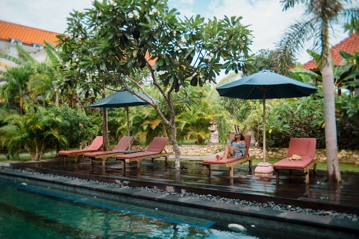 Cozy tropical bedroom with a wooden double bed, orange sheets, patterned pillows, abstract black-and-white wall art, and ornate wall sconces. - Bali Villas