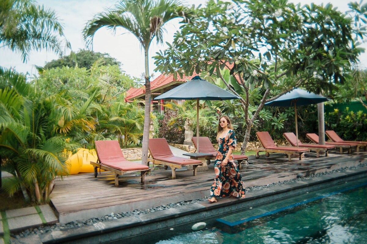Woman in floral dress standing by infinity pool at tropical Bali resort with lounge chairs, palm trees, and thatched umbrellas. - Bali Villas
