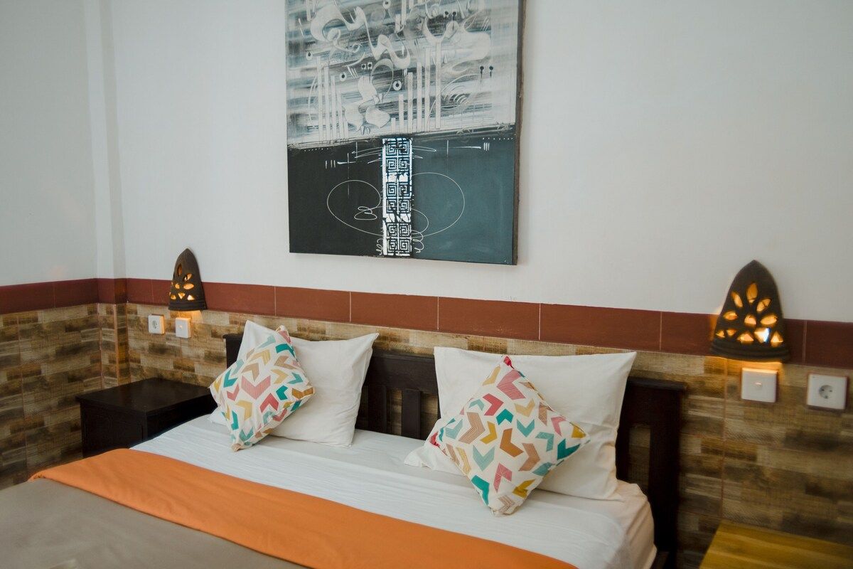 Cozy tropical bedroom with a wooden double bed, orange sheets, patterned pillows, abstract black-and-white wall art, and ornate wall sconces. - Bali Villas