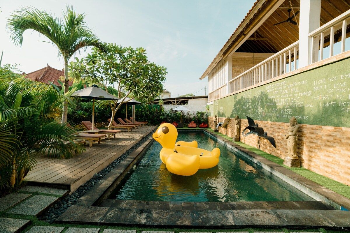 Cozy tropical bedroom with a wooden double bed, orange sheets, patterned pillows, abstract black-and-white wall art, and ornate wall sconces. - Bali Villas