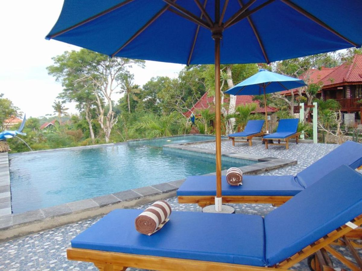 Serene infinity pool at a Bali resort with blue lounge chairs, umbrellas, towels, and lush tropical greenery. - Bali Villas