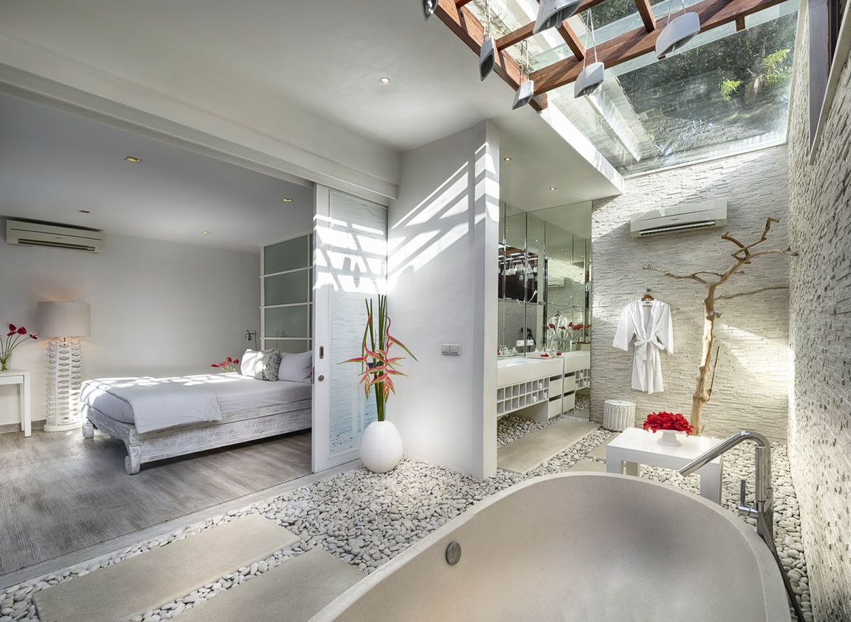 Luxurious spa bathroom with a freestanding tub filled with red rose petals and floating candles, orchids, white towels, and bamboo-shaded tropical window. - Bali Villas