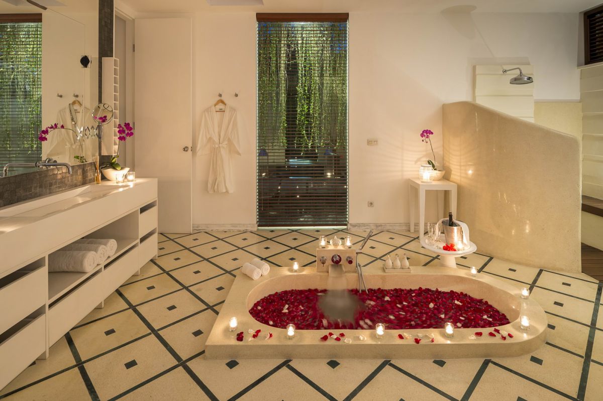 Luxurious spa bathroom with a freestanding tub filled with red rose petals and floating candles, orchids, white towels, and bamboo-shaded tropical window. - Bali Villas
