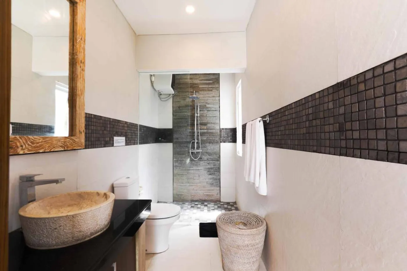Modern bathroom with wooden bowl sink on black vanity, white toilet, glass shower enclosure, and neutral tiled walls. - Bali Villas
