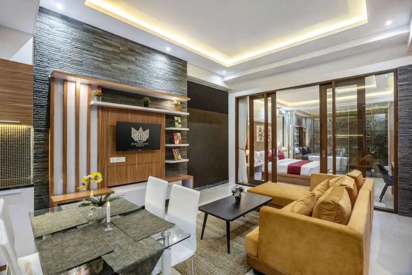 Modern open-plan kitchen and dining area with wooden cabinets, glass table, white chairs, and wall-mounted TV. - Bali Villas