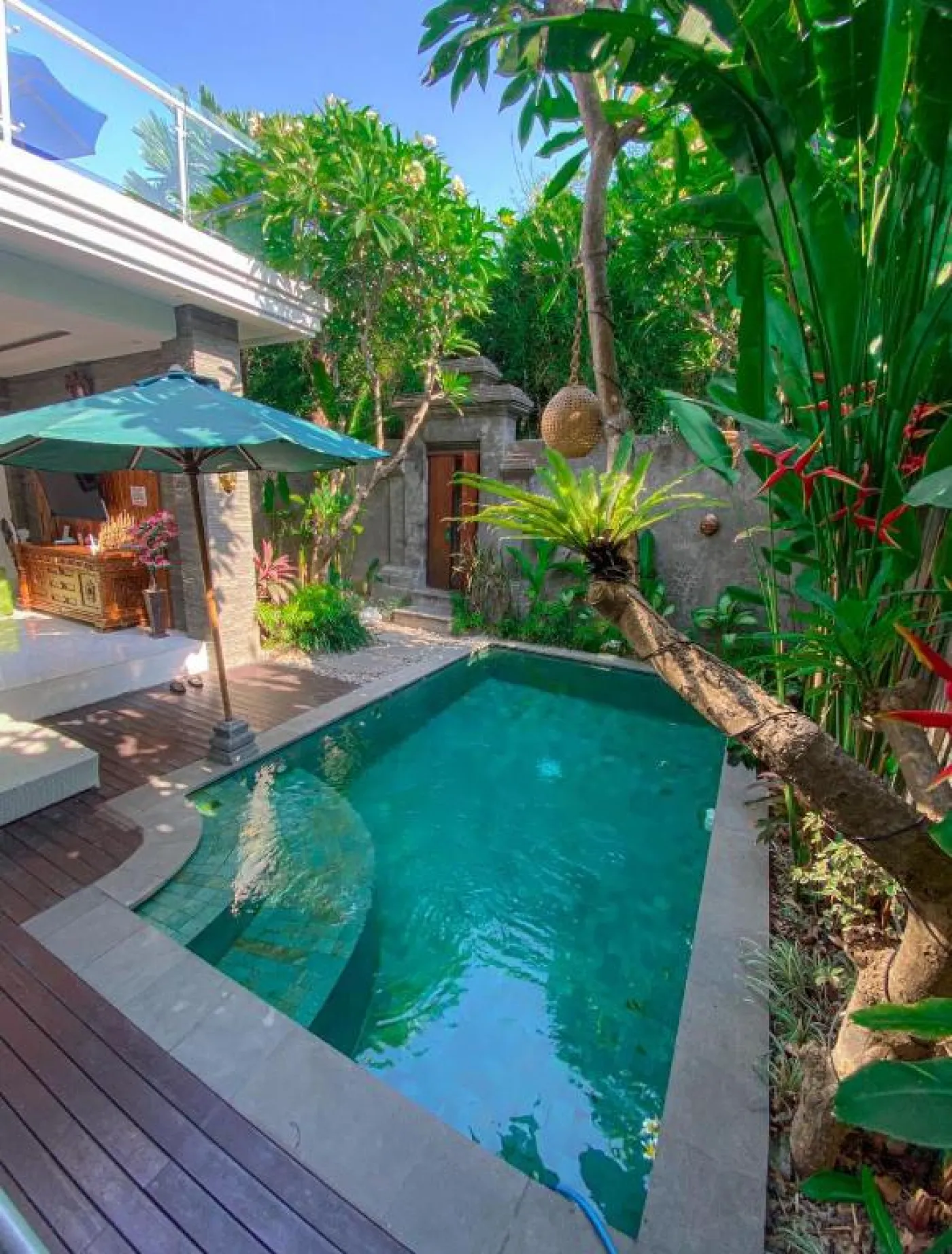 Bright Balinese dining area with wooden table and chairs, framed Hindu goddess artwork, potted plants, and open kitchen under warm ceiling lights. - Bali Villas