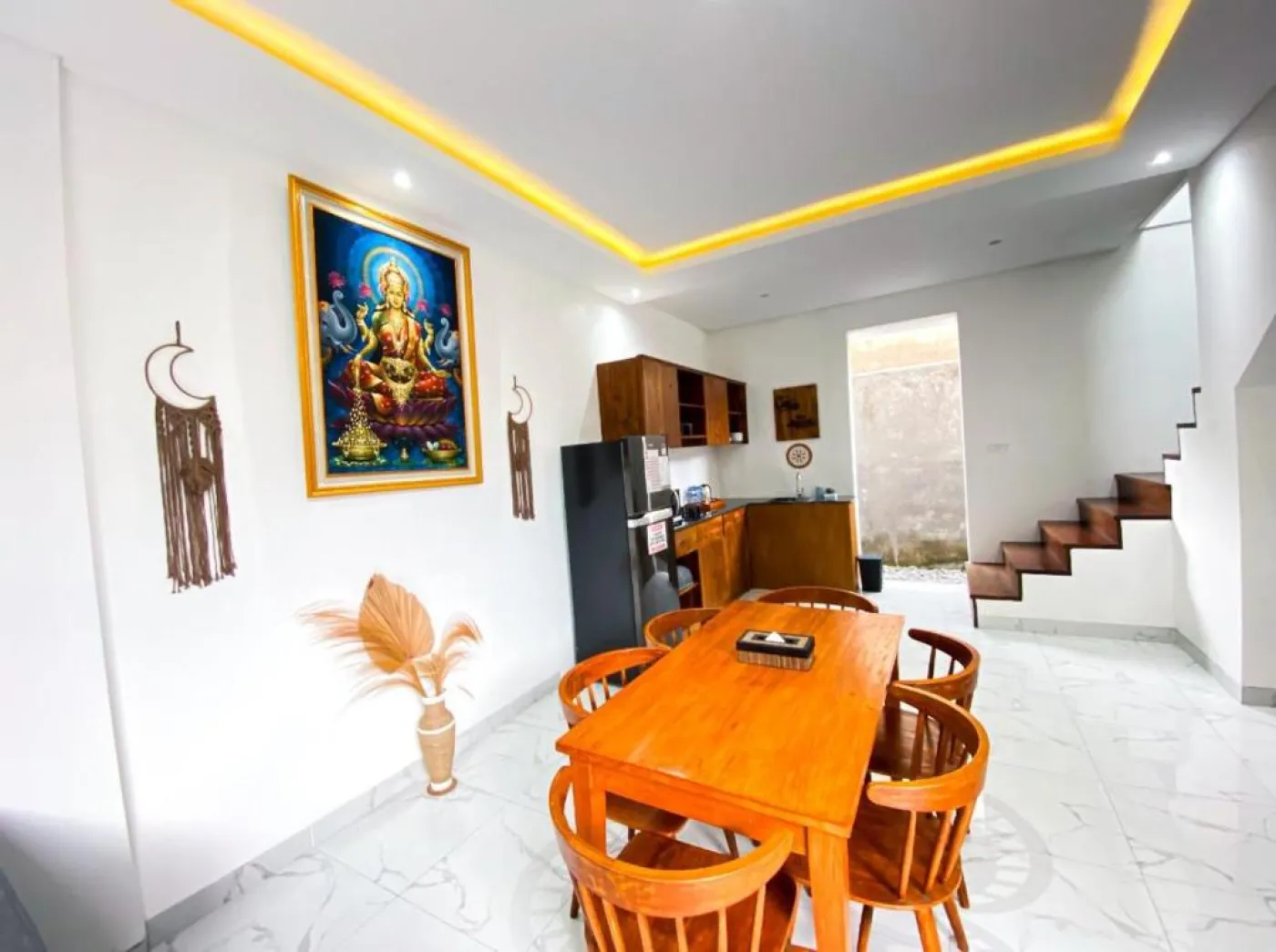 Bright Balinese dining area with wooden table and chairs, framed Hindu goddess artwork, potted plants, and open kitchen under warm ceiling lights. - Bali Villas