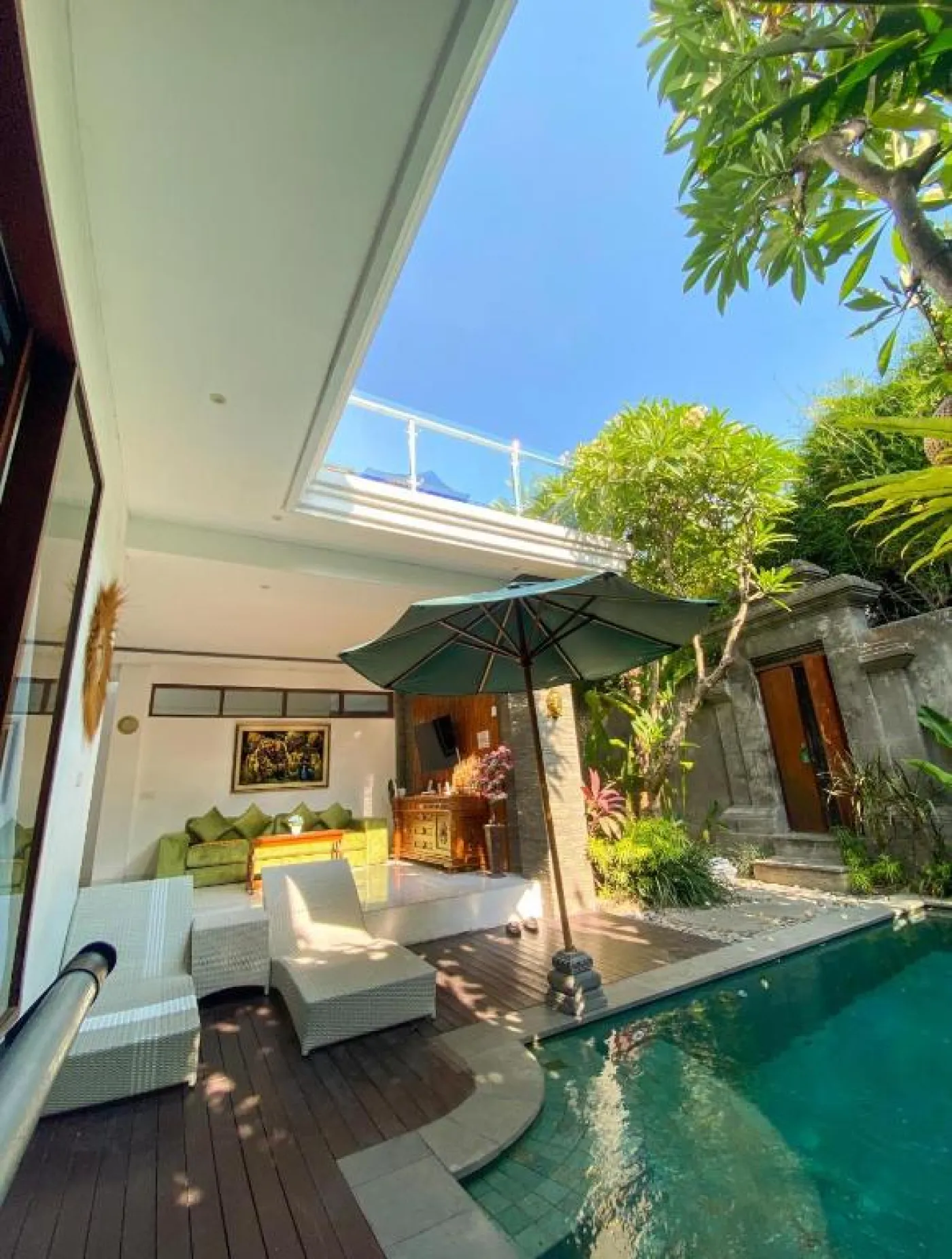 Bright Balinese dining area with wooden table and chairs, framed Hindu goddess artwork, potted plants, and open kitchen under warm ceiling lights. - Bali Villas