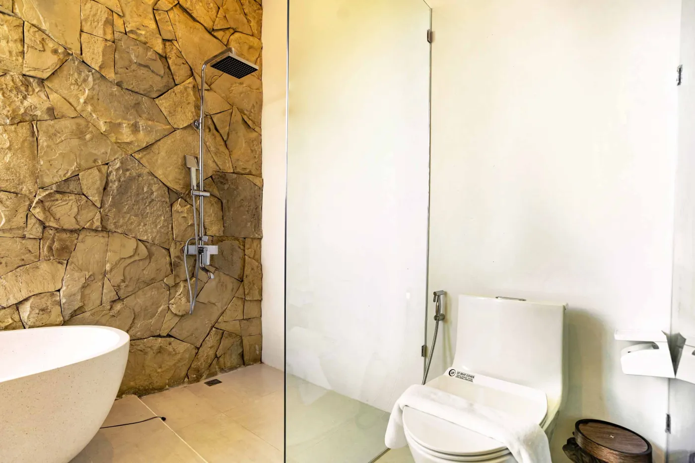 Luxurious modern bathroom with textured stone walls, arched black-framed windows overlooking tropical palms, freestanding white bathtub filled with red petals, and sleek vanity sink. - Bali Villas