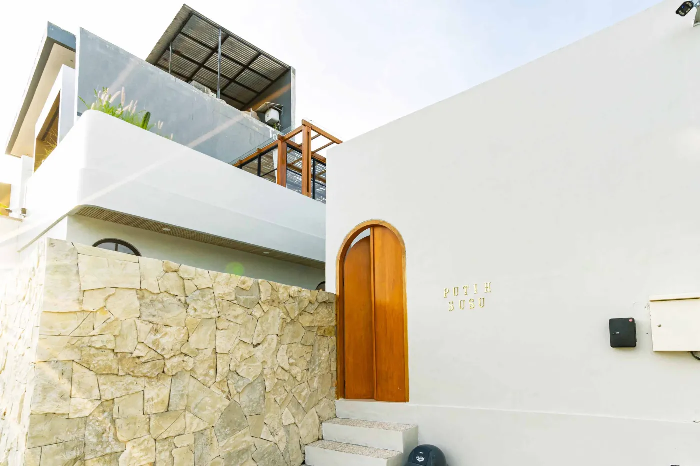 Luxurious modern bathroom with textured stone walls, arched black-framed windows overlooking tropical palms, freestanding white bathtub filled with red petals, and sleek vanity sink. - Bali Villas