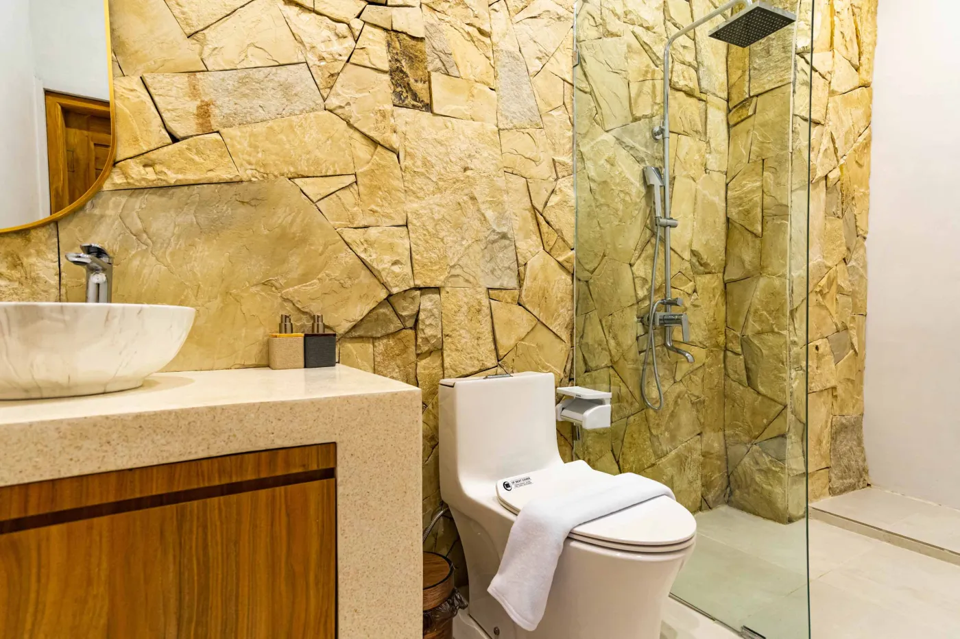 Luxurious modern bathroom with textured stone walls, arched black-framed windows overlooking tropical palms, freestanding white bathtub filled with red petals, and sleek vanity sink. - Bali Villas