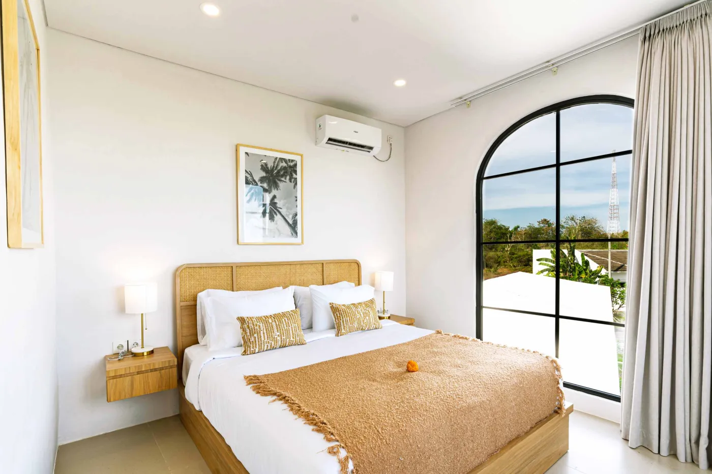Luxurious modern bathroom with textured stone walls, arched black-framed windows overlooking tropical palms, freestanding white bathtub filled with red petals, and sleek vanity sink. - Bali Villas