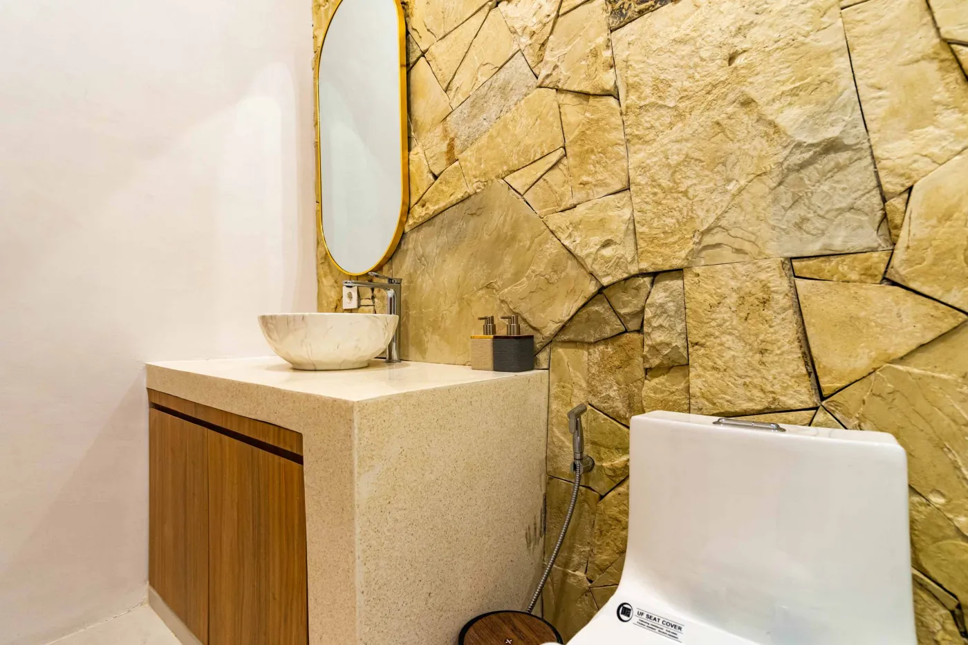 Luxurious modern bathroom with textured stone walls, arched black-framed windows overlooking tropical palms, freestanding white bathtub filled with red petals, and sleek vanity sink. - Bali Villas