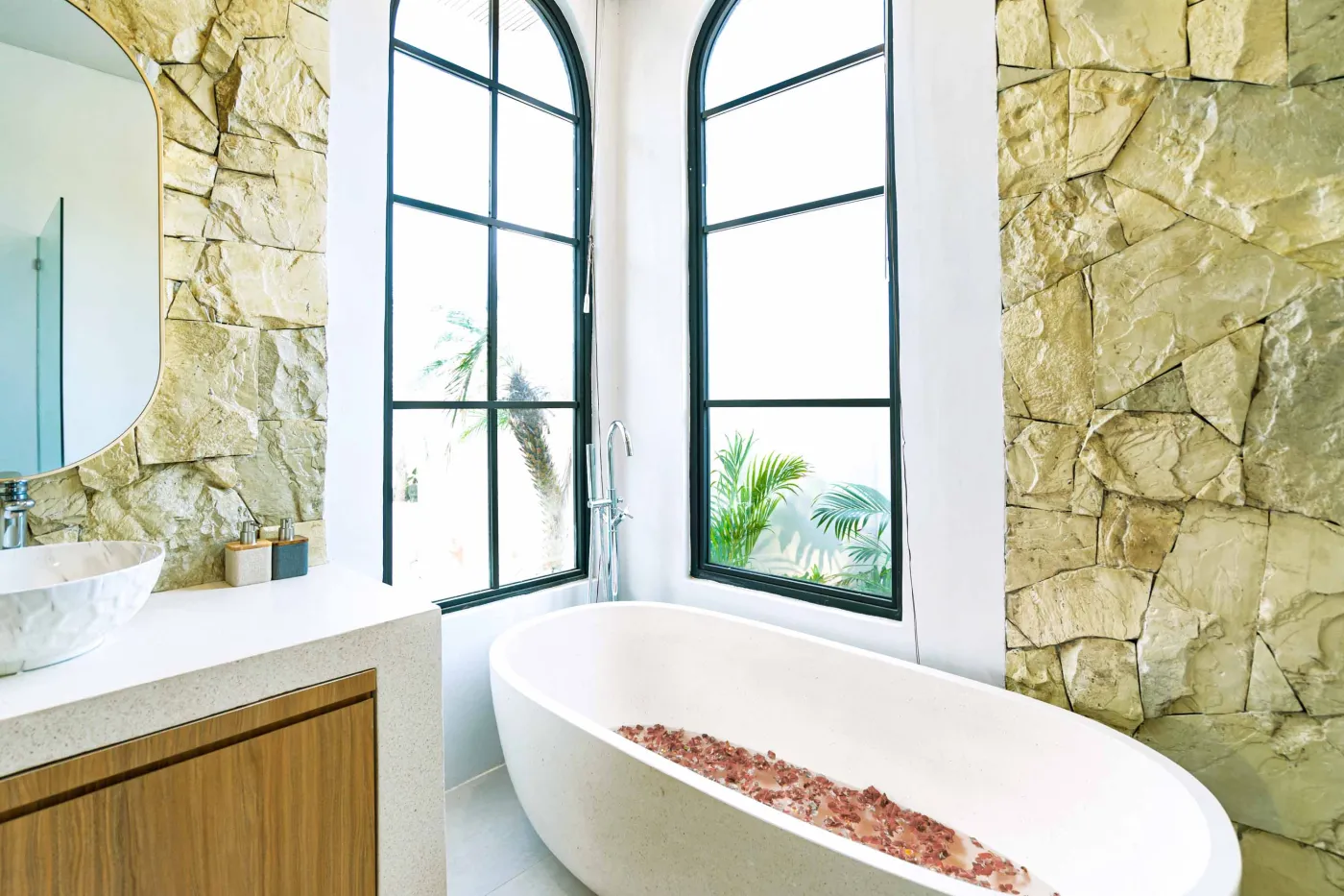Luxurious modern bathroom with textured stone walls, arched black-framed windows overlooking tropical palms, freestanding white bathtub filled with red petals, and sleek vanity sink. - Bali Villas