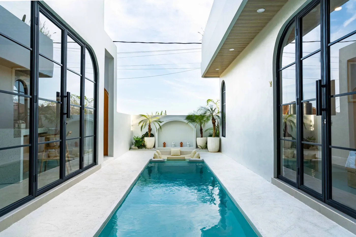 Modern minimalist courtyard featuring a narrow turquoise infinity pool, white arched architecture, potted palms, and cream lounge seating. - Bali Villas