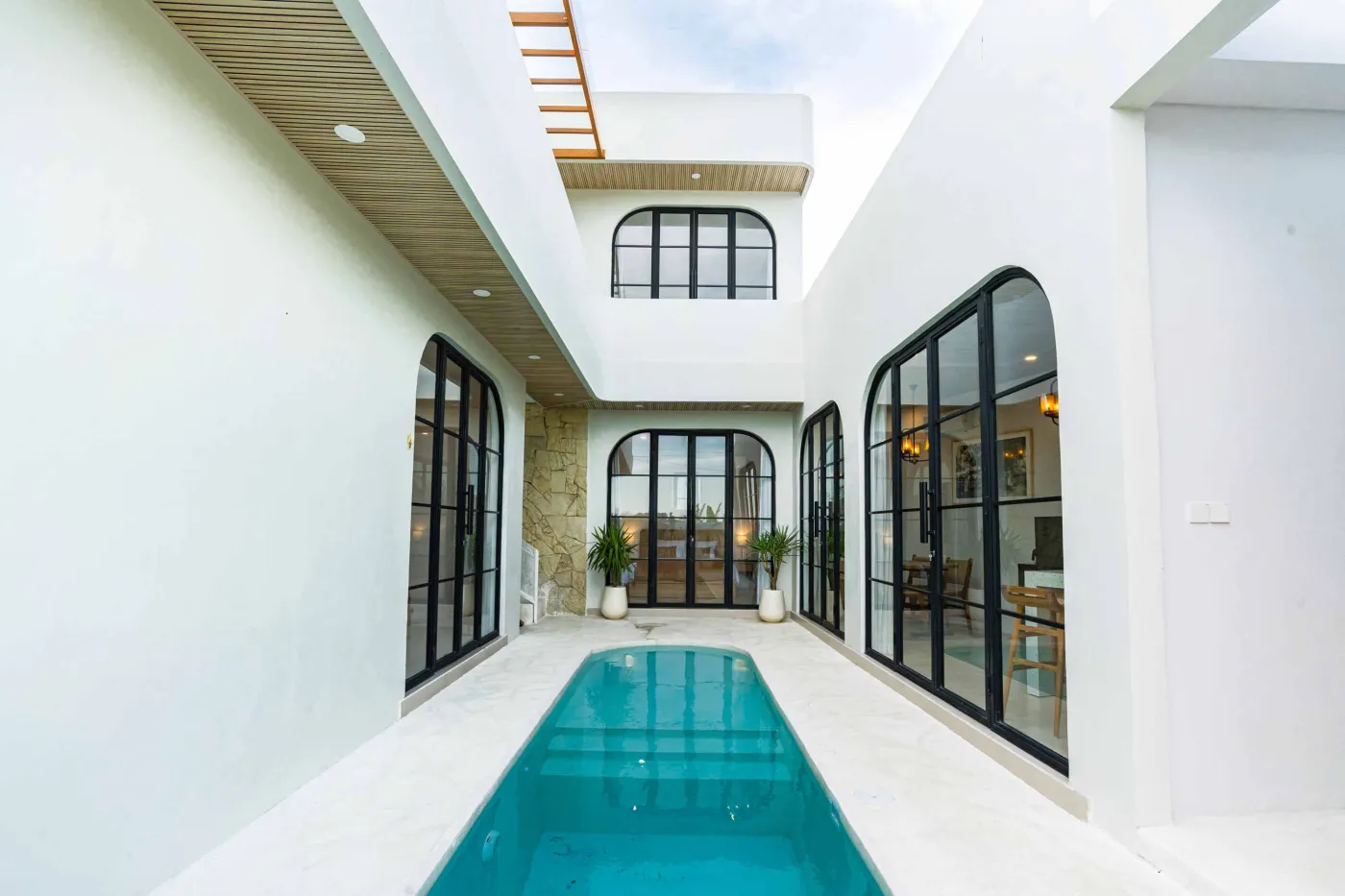 Luxurious modern bathroom with textured stone walls, arched black-framed windows overlooking tropical palms, freestanding white bathtub filled with red petals, and sleek vanity sink. - Bali Villas