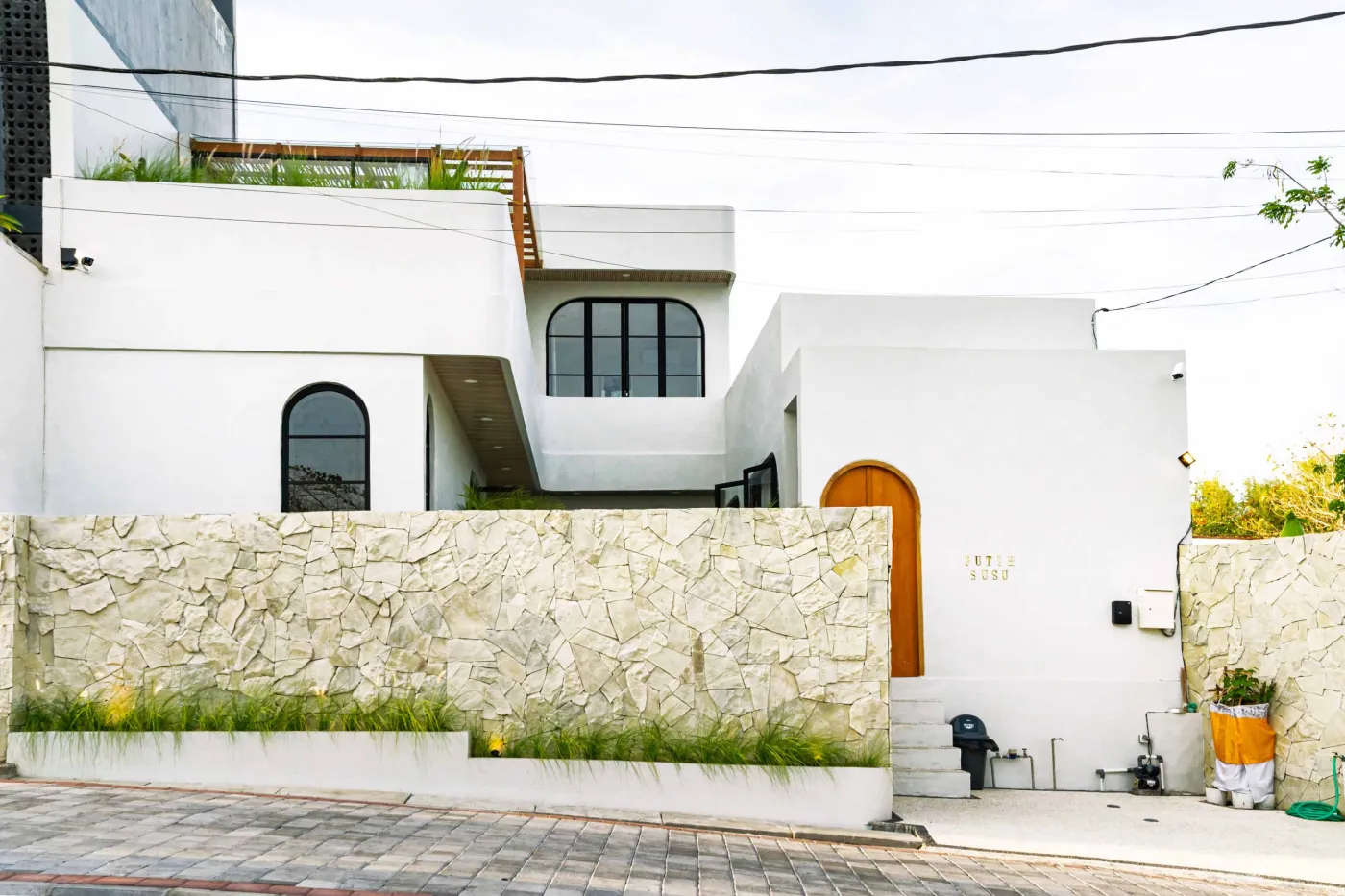 Luxurious modern bathroom with textured stone walls, arched black-framed windows overlooking tropical palms, freestanding white bathtub filled with red petals, and sleek vanity sink. - Bali Villas