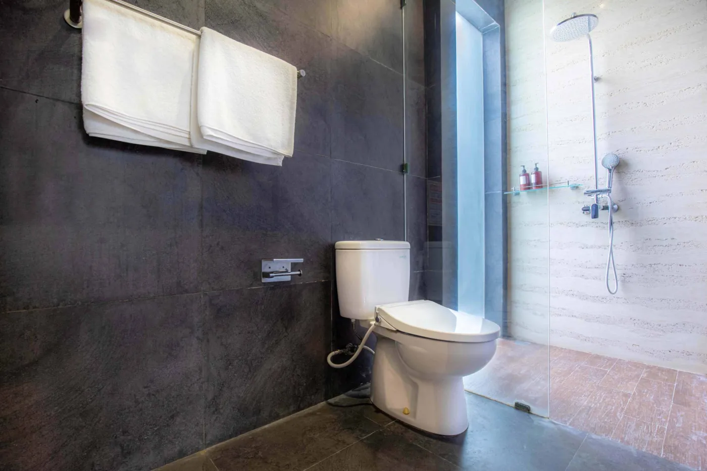 Modern black-tiled bathroom with freestanding white bathtub, handheld shower, and folded towels on rack. - Bali Villas