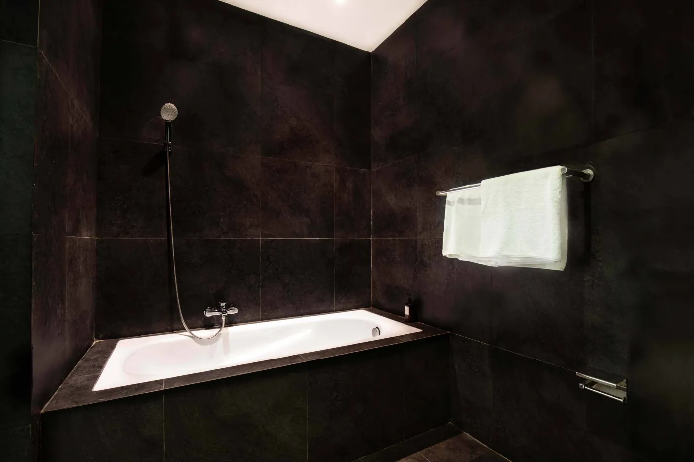 Modern black-tiled bathroom with freestanding white bathtub, handheld shower, and folded towels on rack. - Bali Villas