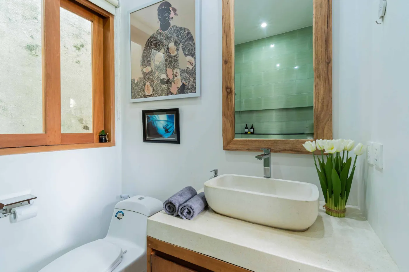 Bright modern Balinese kitchen with white countertops, wooden cabinets, potted plants, large windows, and a wall-mounted TV. - Bali Villas