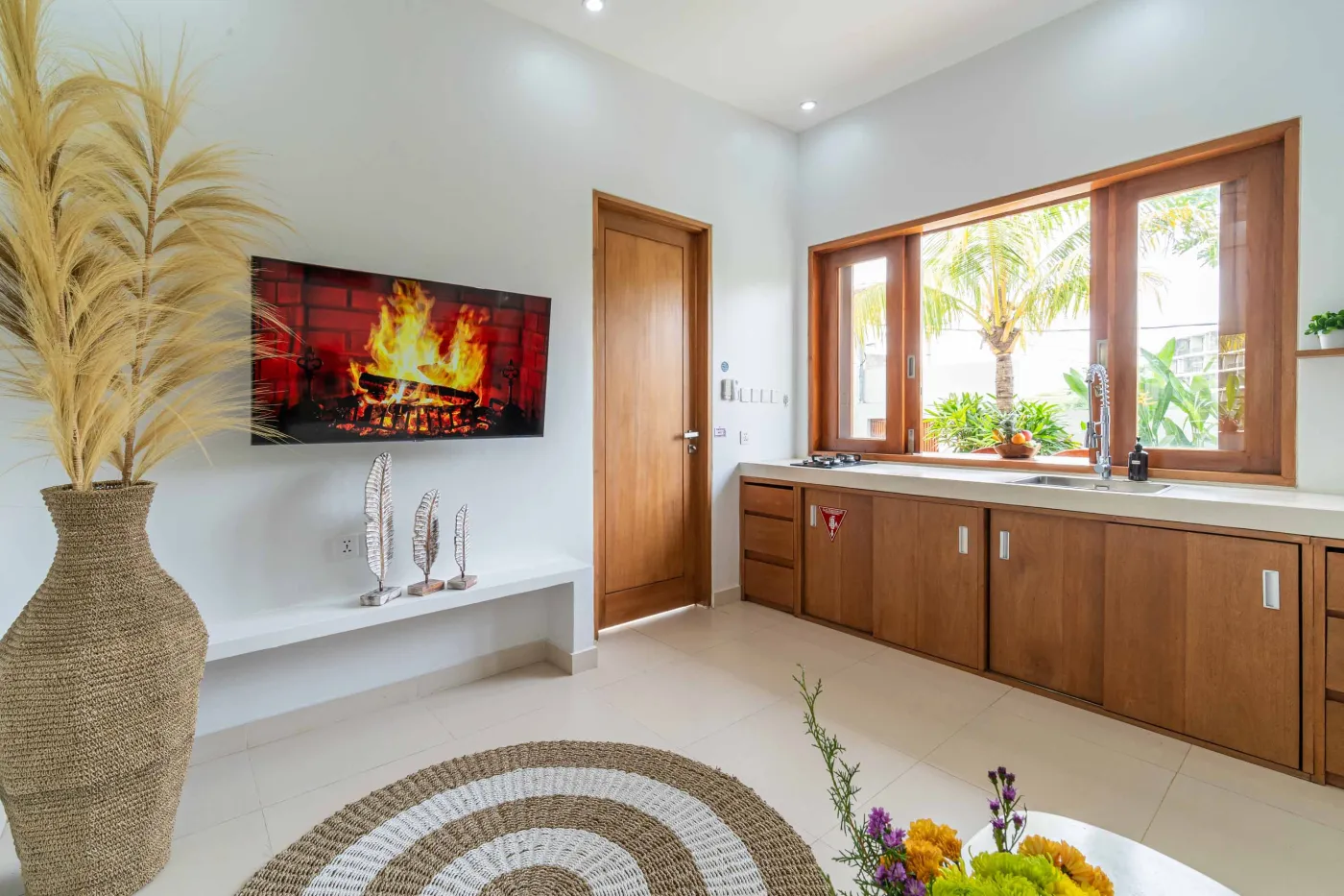 Bright modern Balinese kitchen with white countertops, wooden cabinets, potted plants, large windows, and a wall-mounted TV. - Bali Villas