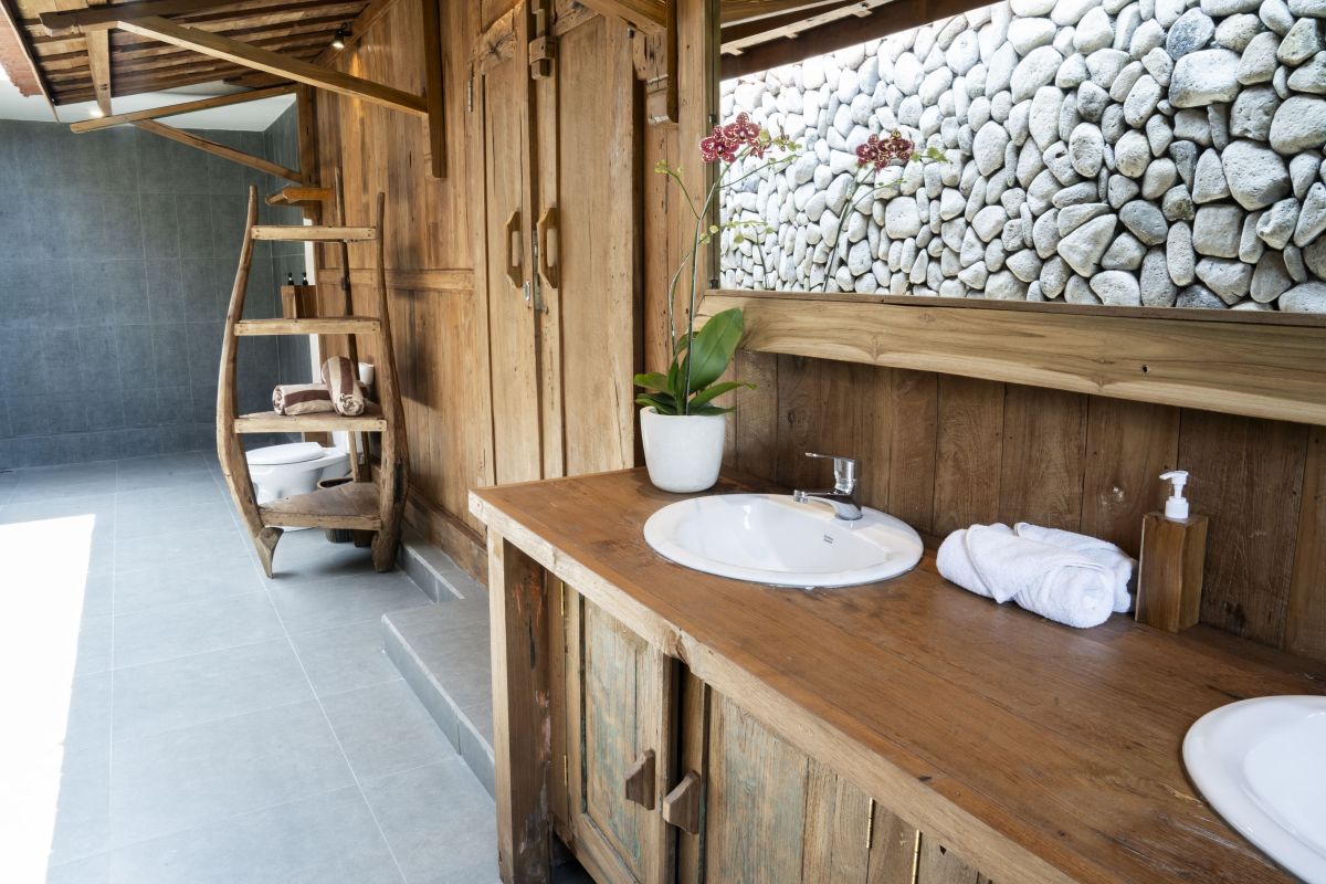 Rustic Balinese bathroom with wooden vanity, double sinks, orchid plant, towels, and pebble stone wall. - Bali Villas