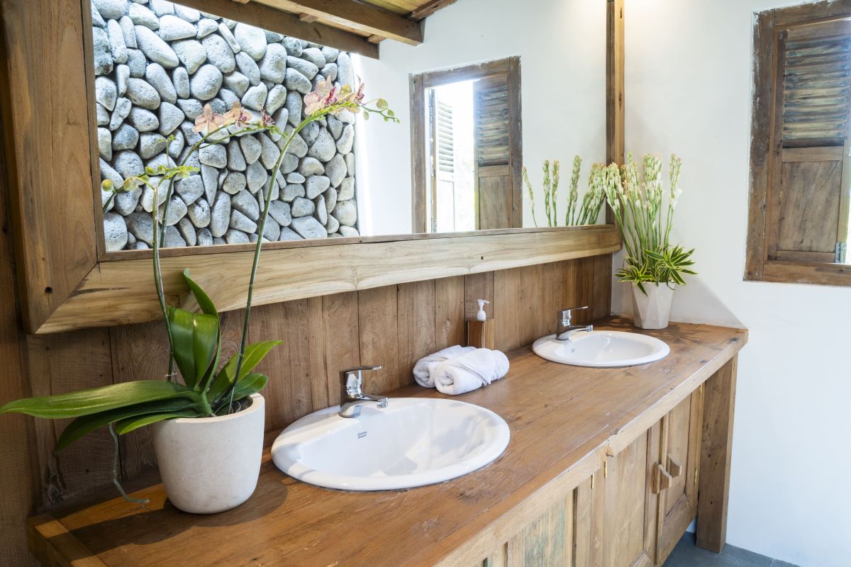 Rustic Balinese bathroom with wooden vanity, double sinks, orchid plant, towels, and pebble stone wall. - Bali Villas