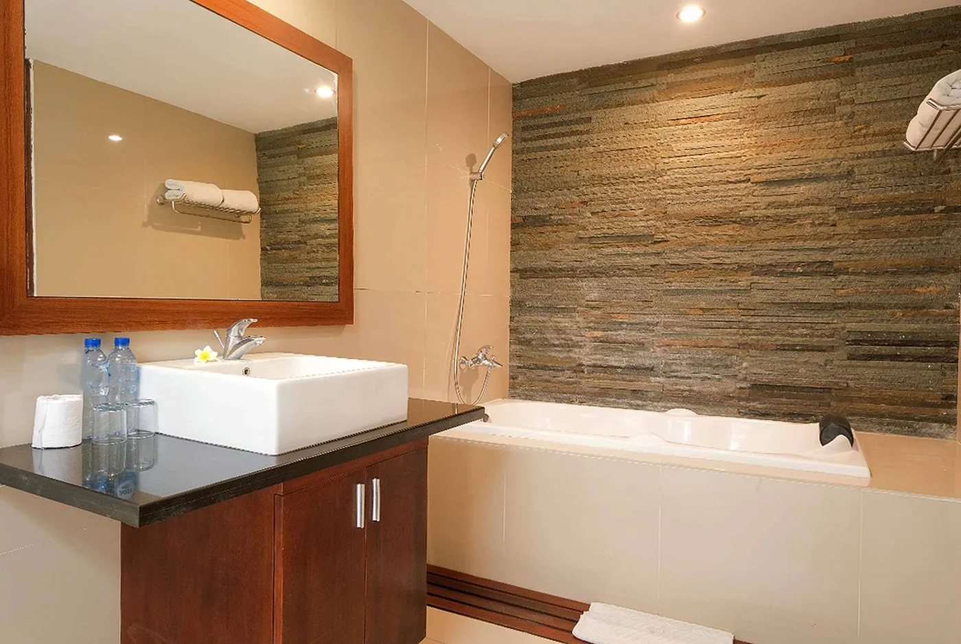 Modern luxury hotel bathroom with wooden vanity, white sink, stone wall, glass shower enclosure, freestanding bathtub, and folded towels. - Bali Villas