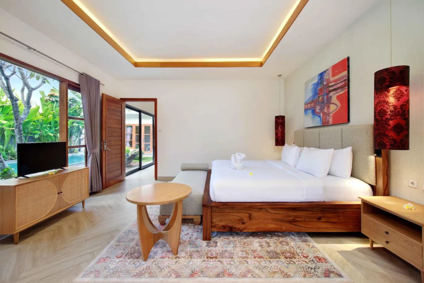 Spacious Balinese-style hotel bedroom with twin wooden platform beds, abstract colorful wall art, mirrored wardrobe, and arched bathroom doorway. - Bali Villas
