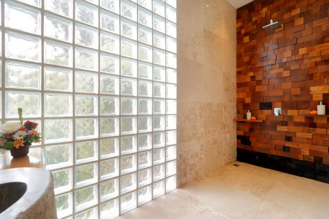 Modern luxury bathroom featuring a frosted glass block window, wooden accent wall with rain showerhead, and fresh flowers on a side table. - Bali Villas