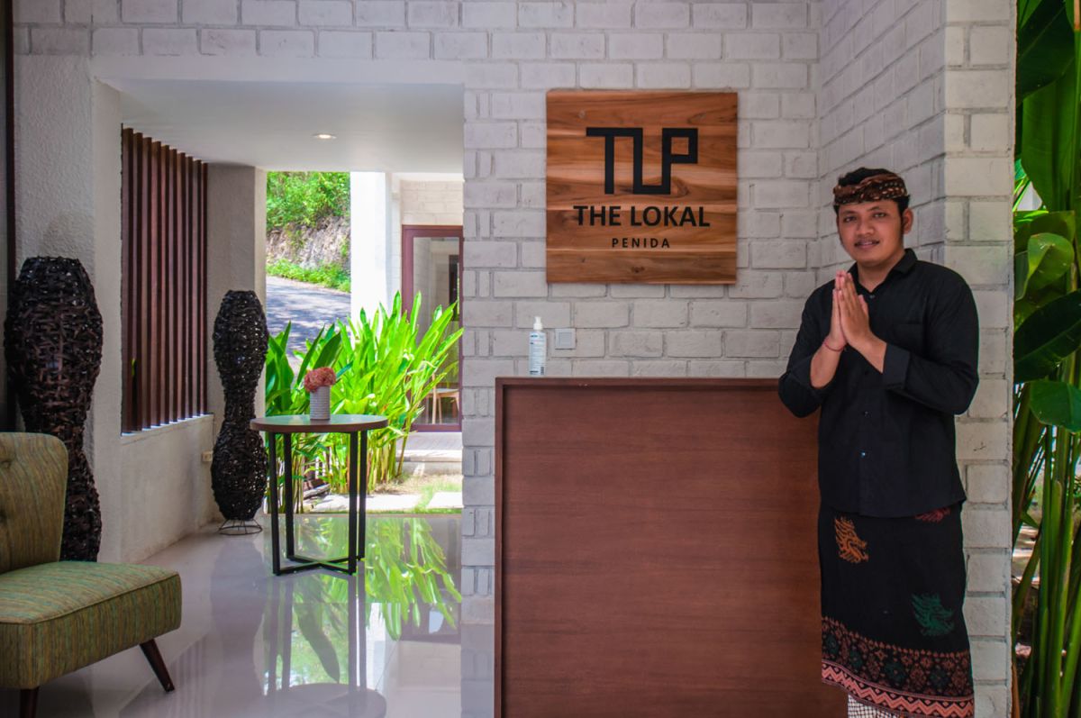 Reception at The Lokale Residences in Bali: smiling Balinese staff in traditional attire greets visitors behind wooden desk amid tropical plants and open courtyard. - Bali Villas