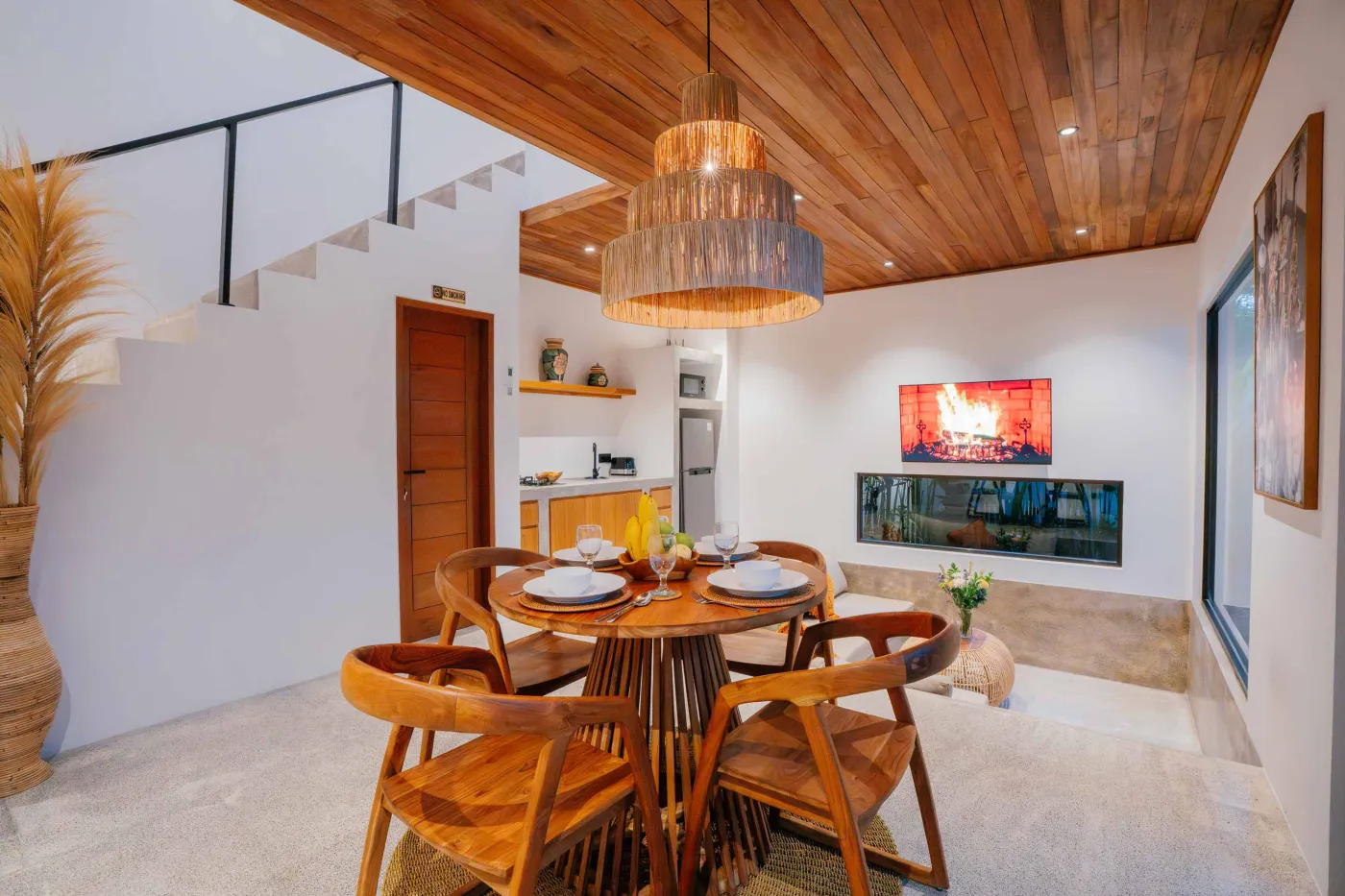 Modern tropical villa living room with low white sofa, rattan coffee table, wall-mounted TV, open kitchen, and floor-to-ceiling windows to infinity pool amid lush greenery. - Bali Villas