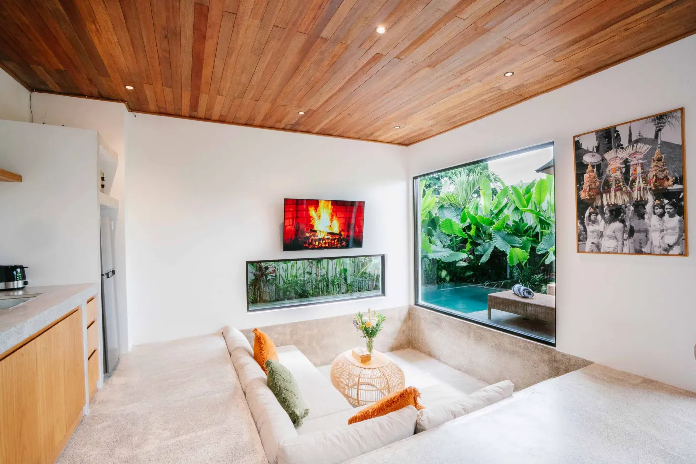 Modern tropical villa living room with low white sofa, rattan coffee table, wall-mounted TV, open kitchen, and floor-to-ceiling windows to infinity pool amid lush greenery. - Bali Villas