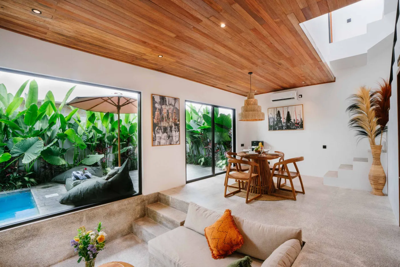 Modern tropical villa living room with low white sofa, rattan coffee table, wall-mounted TV, open kitchen, and floor-to-ceiling windows to infinity pool amid lush greenery. - Bali Villas
