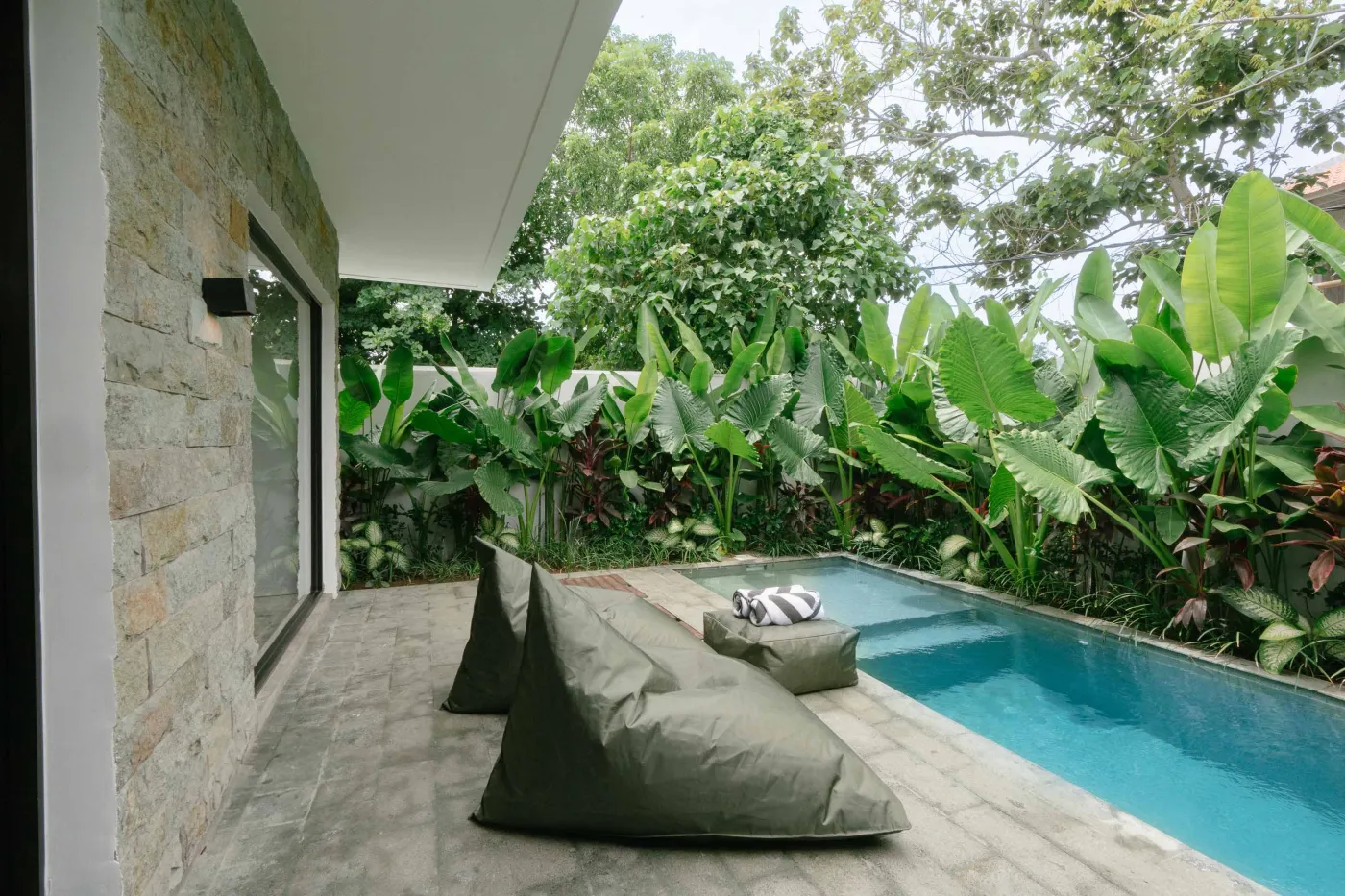 Modern tropical villa living room with low white sofa, rattan coffee table, wall-mounted TV, open kitchen, and floor-to-ceiling windows to infinity pool amid lush greenery. - Bali Villas