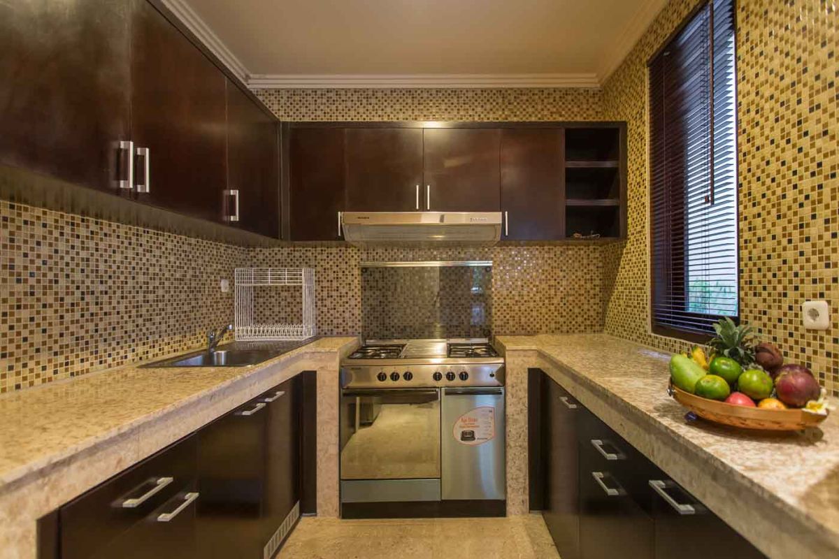 Modern U-shaped kitchen with dark wood cabinets, gold mosaic tile backsplash, granite counters, gas stove, and a bowl of fresh tropical fruits. - Bali Villas