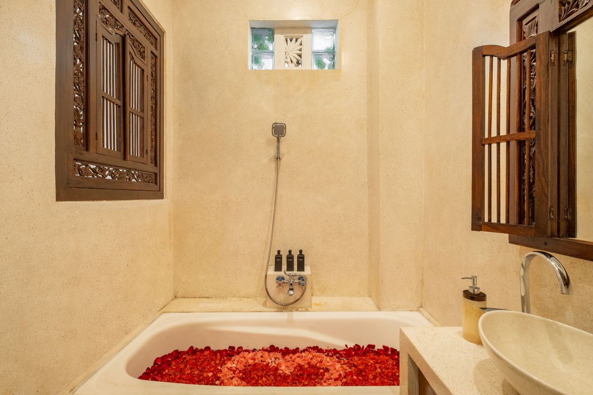 Elegant Balinese-style bathroom with white bathtub filled with red rose petals, wooden carved shutters, wall-mounted shower, and sleek sink. - Bali Villas