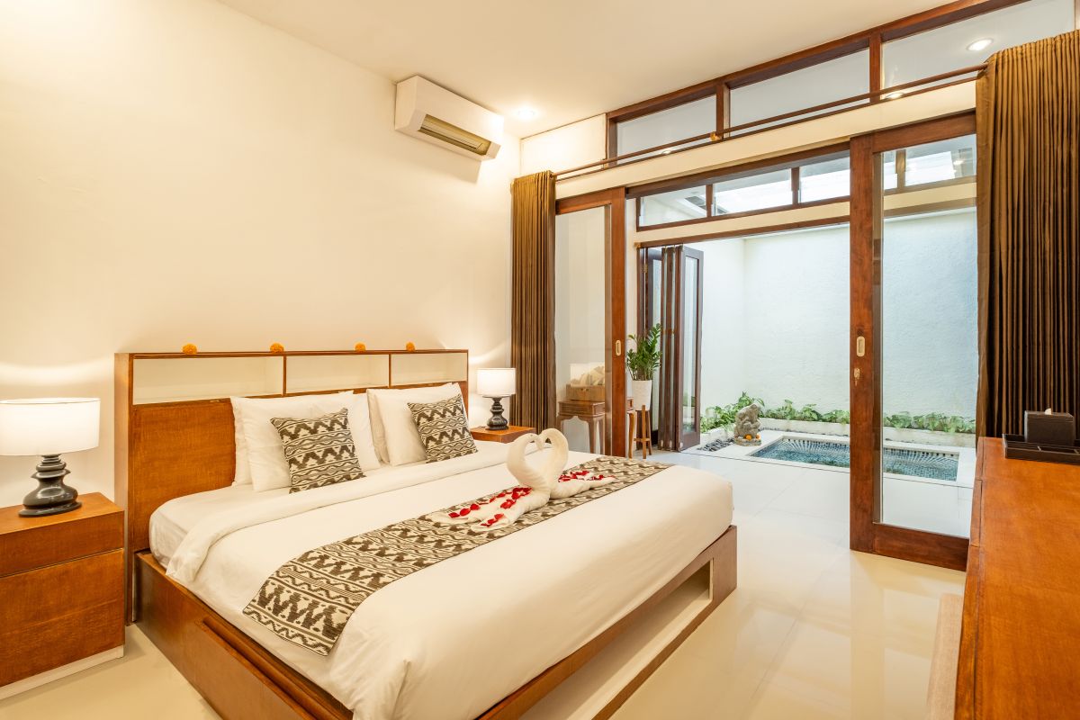 Elegant Balinese-style bathroom with white bathtub filled with red rose petals, wooden carved shutters, wall-mounted shower, and sleek sink. - Bali Villas