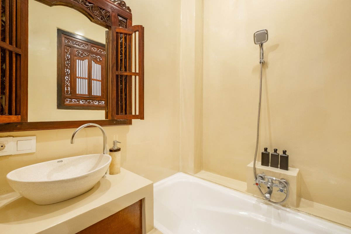 Elegant Balinese-style bathroom with white bathtub filled with red rose petals, wooden carved shutters, wall-mounted shower, and sleek sink. - Bali Villas