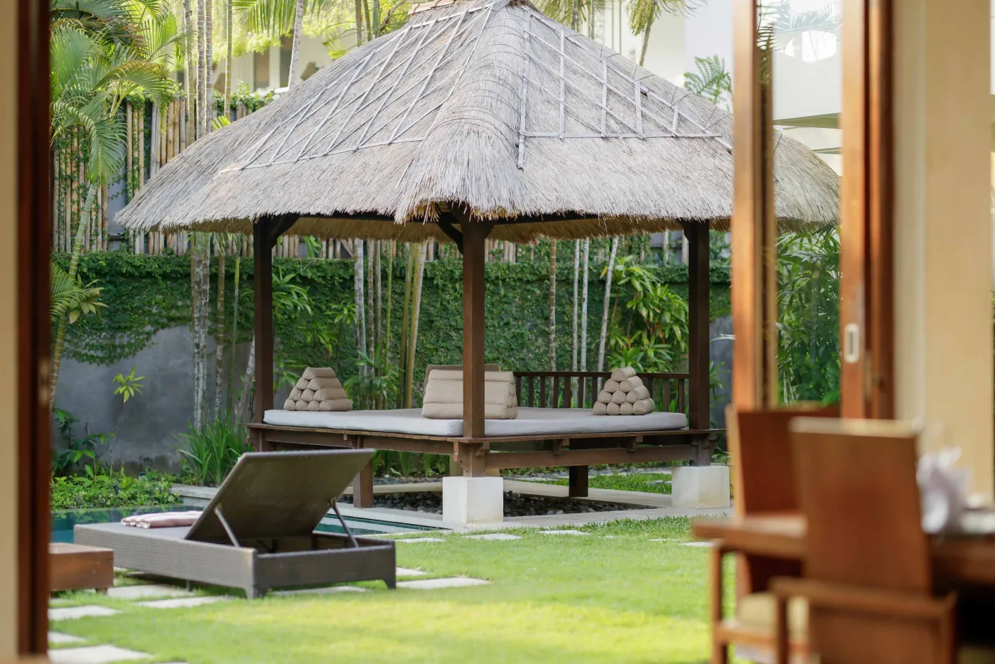 Traditional Balinese thatched pavilion with cushioned daybed in lush tropical garden, lounge chair by small pool. - Bali Villas