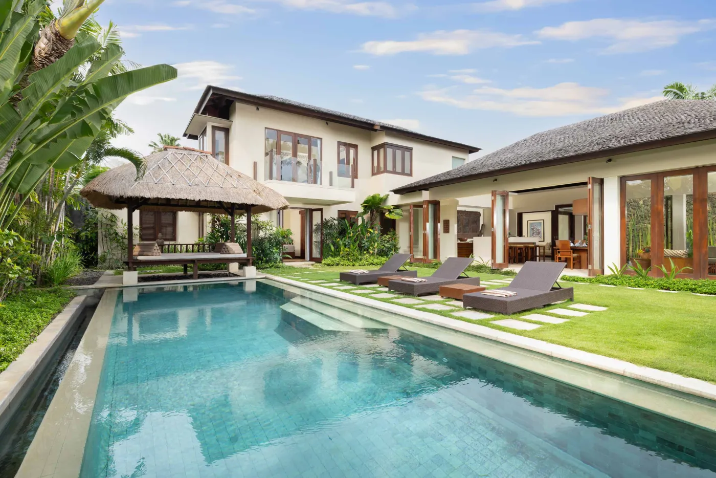 Luxurious Balinese villa with turquoise infinity pool, tropical gardens, and lounge seating under clear skies. - Bali Villas