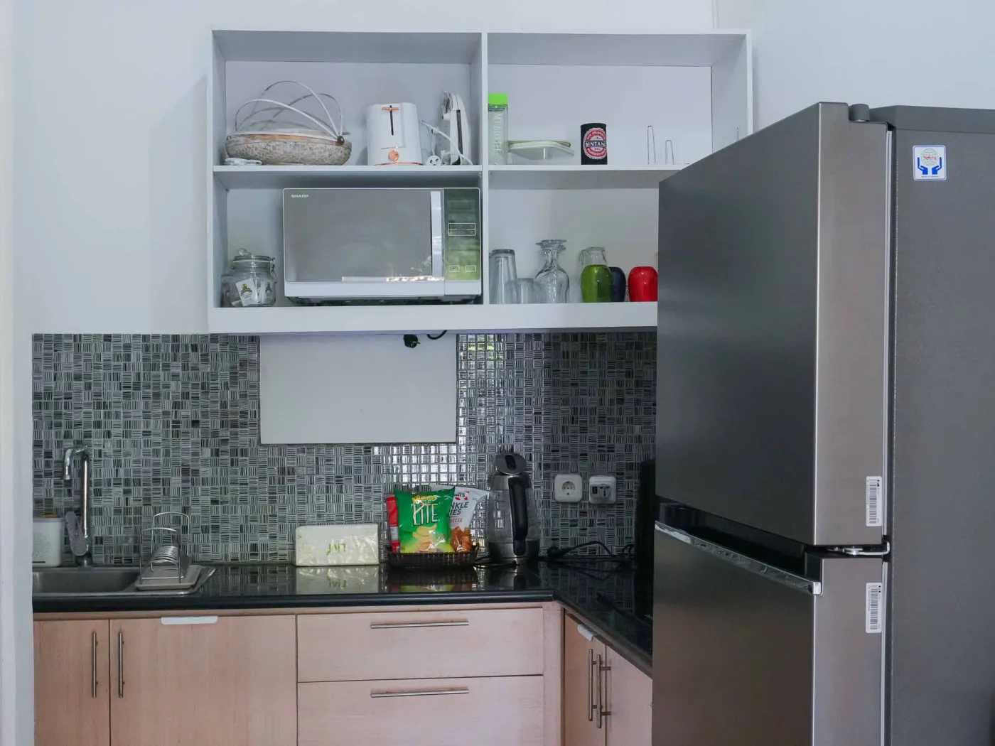 Modern compact kitchen featuring light wooden cabinets, black countertop, sink, microwave, toaster, and green mosaic backsplash. - Bali Villas