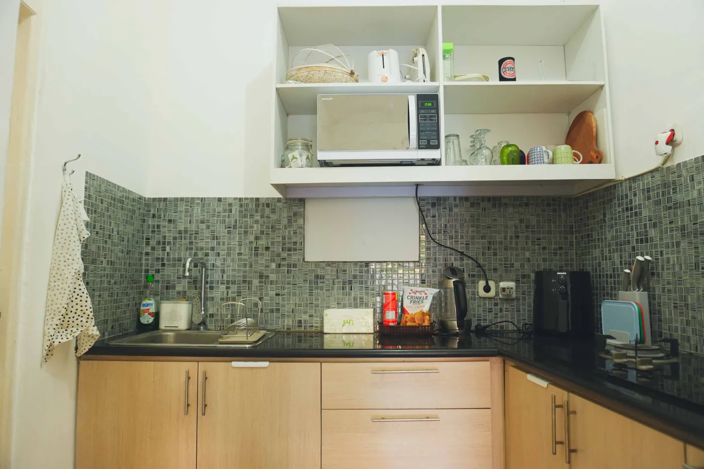 Modern compact kitchen featuring light wooden cabinets, black countertop, sink, microwave, toaster, and green mosaic backsplash. - Bali Villas