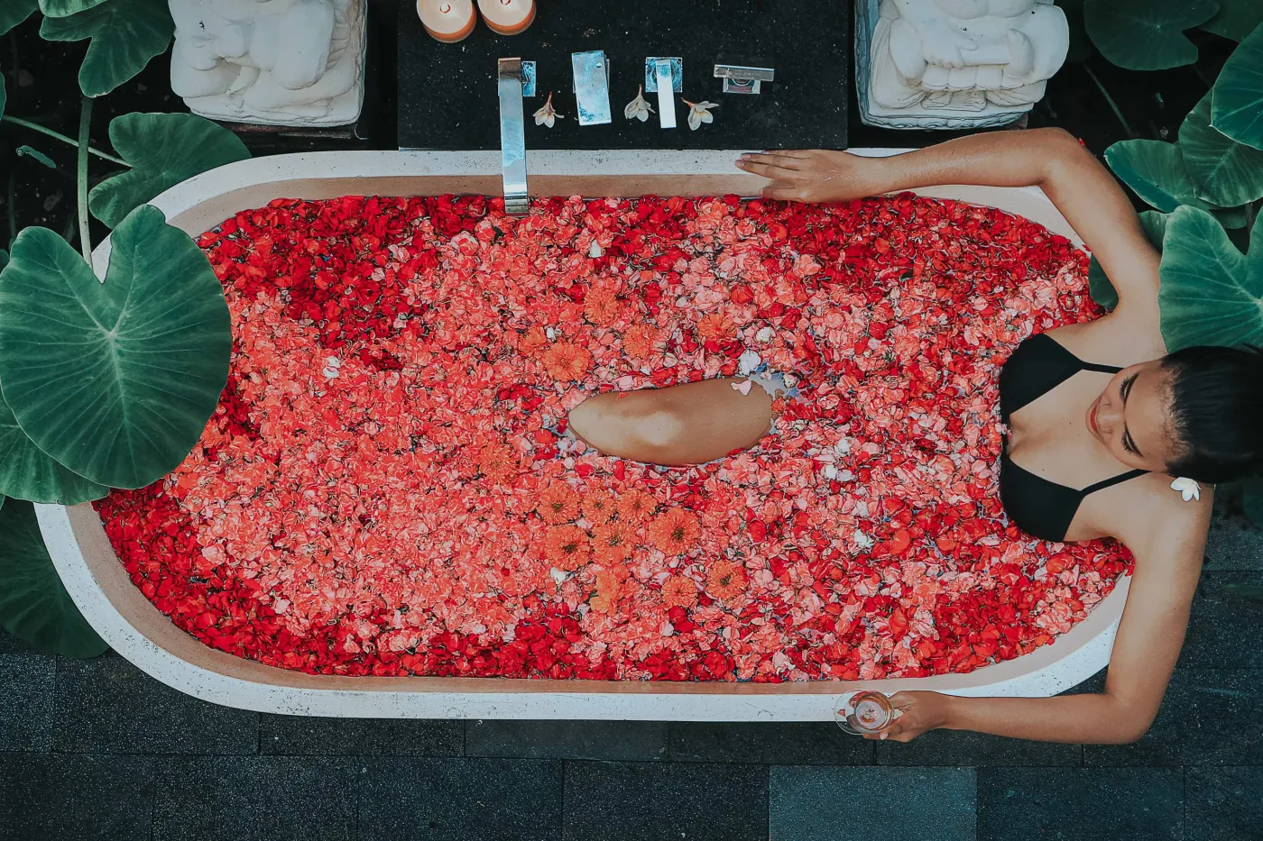 Overhead view of a woman relaxing in a clawfoot bathtub filled with red rose petals, surrounded by tropical plants, Buddha statues, and spa amenities. - Bali Villas