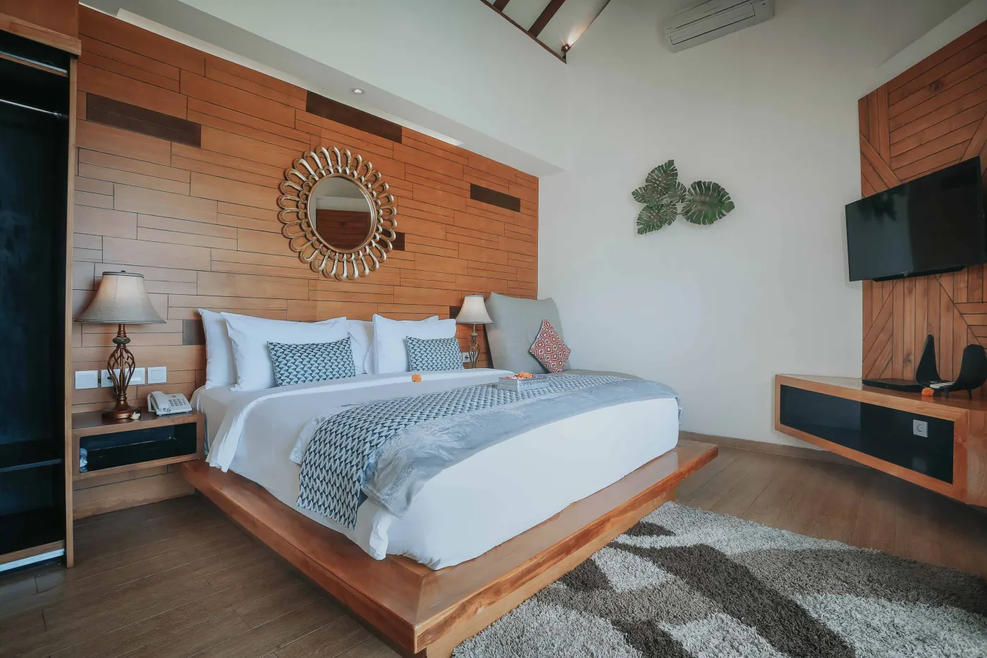 Luxurious Bali villa bedroom with king bed, wooden accents, wall mirror, and open balcony doors overlooking infinity pool and tropical palms. - Bali Villas