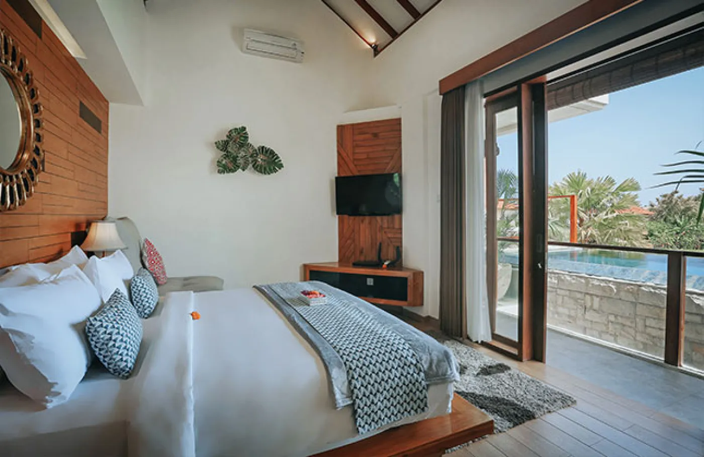 Luxurious Bali villa bedroom with king bed, wooden accents, wall mirror, and open balcony doors overlooking infinity pool and tropical palms. - Bali Villas
