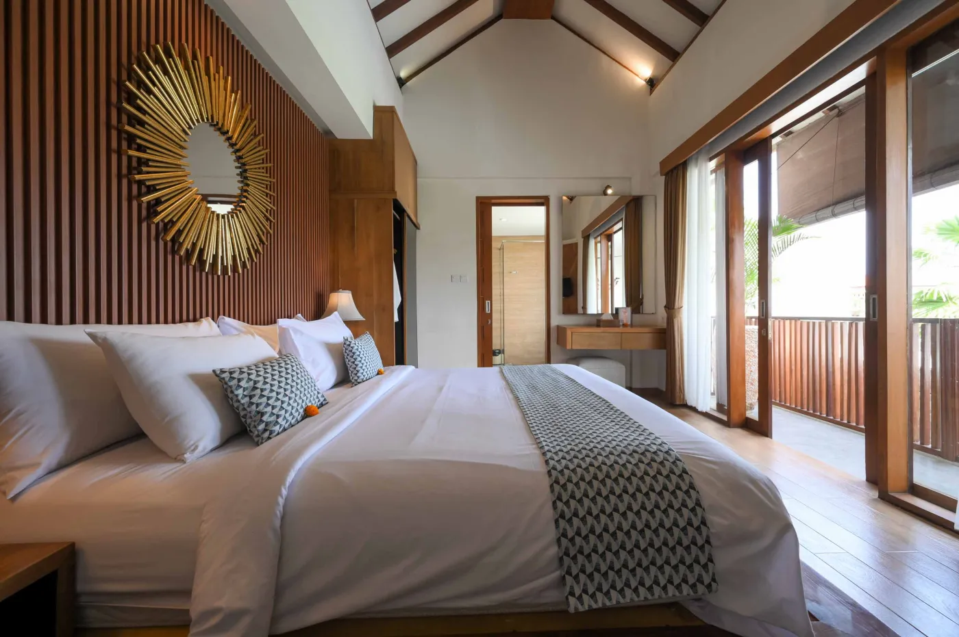 Luxurious Bali villa bedroom with king bed, wooden accents, wall mirror, and open balcony doors overlooking infinity pool and tropical palms. - Bali Villas