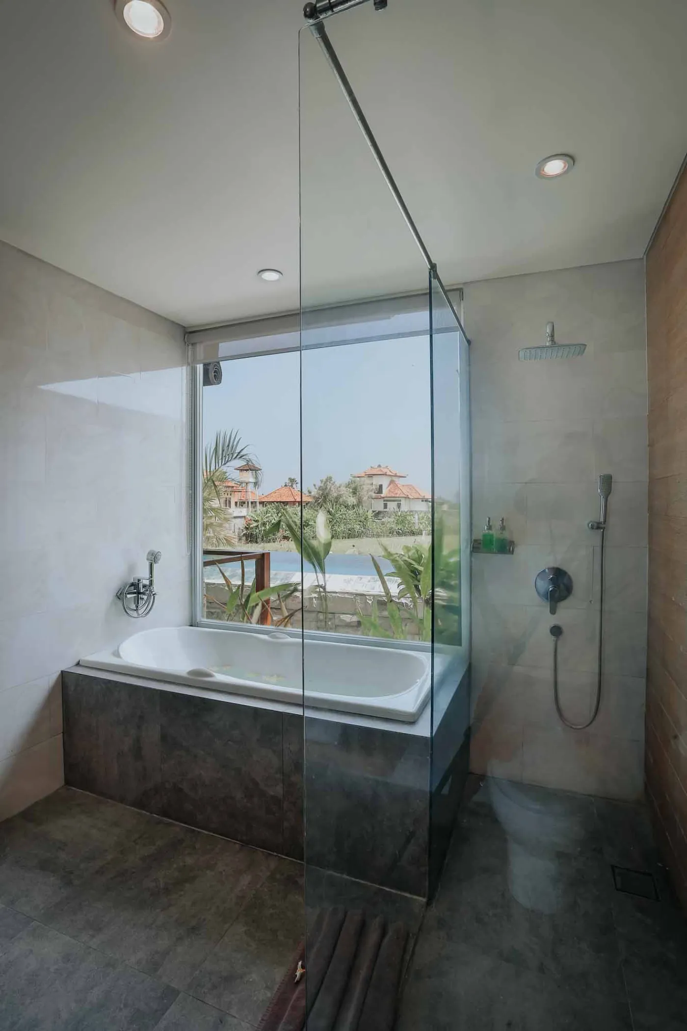 Modern luxury bathroom with freestanding white bathtub, glass shower enclosure, and floor-to-ceiling window overlooking tropical palm trees, pool, and villas. - Bali Villas