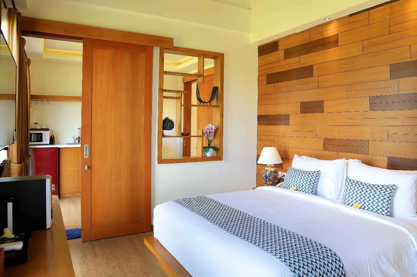 Spacious tropical villa bedroom with wooden walls, white king bed, en-suite bathtub, and wall-mounted TV. - Bali Villas