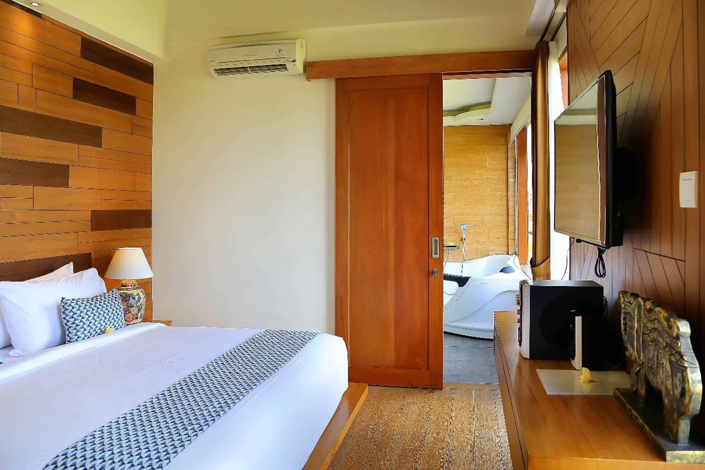 Spacious tropical villa bedroom with wooden walls, white king bed, en-suite bathtub, and wall-mounted TV. - Bali Villas